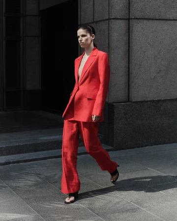 Fashion shot of a young adult woman wearing a red suit, walking outdoors on a city street with a gray building in the background.