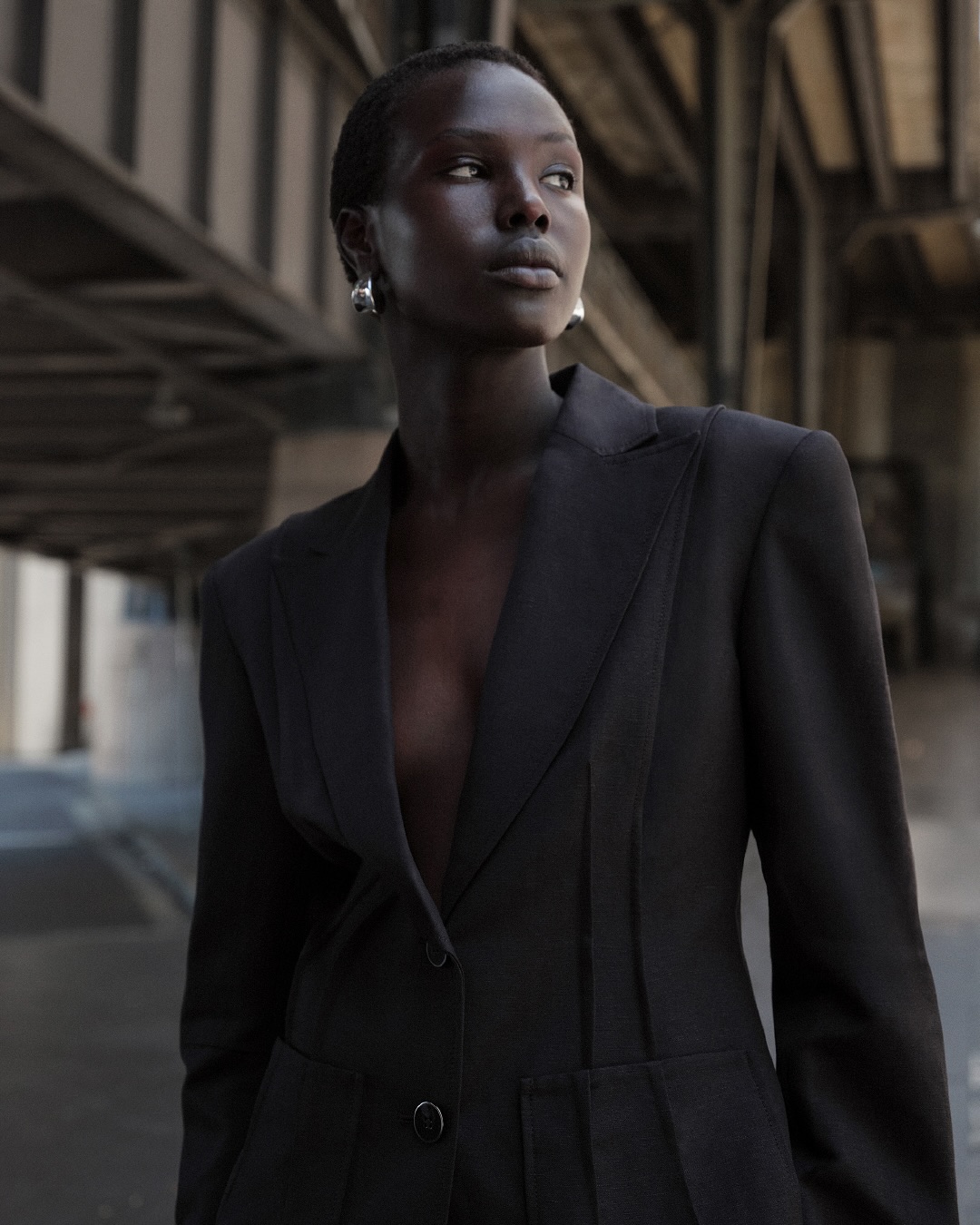 Dark-skinned woman in a black blazer stands under a metal structure in a close-up shot with shallow depth of field, fashion editorial.