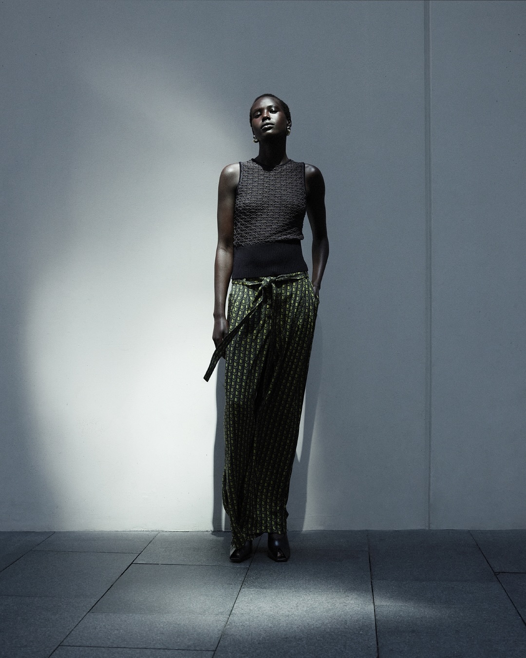An African-American woman stands in front of a textured wall in green, black, and brown patterned pants, a sleeveless top, and black shoes.