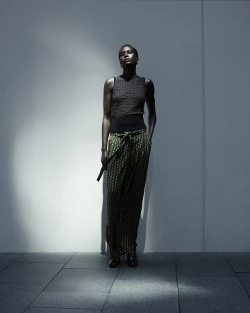 An African-American woman stands in front of a textured wall in green, black, and brown patterned pants, a sleeveless top, and black shoes.