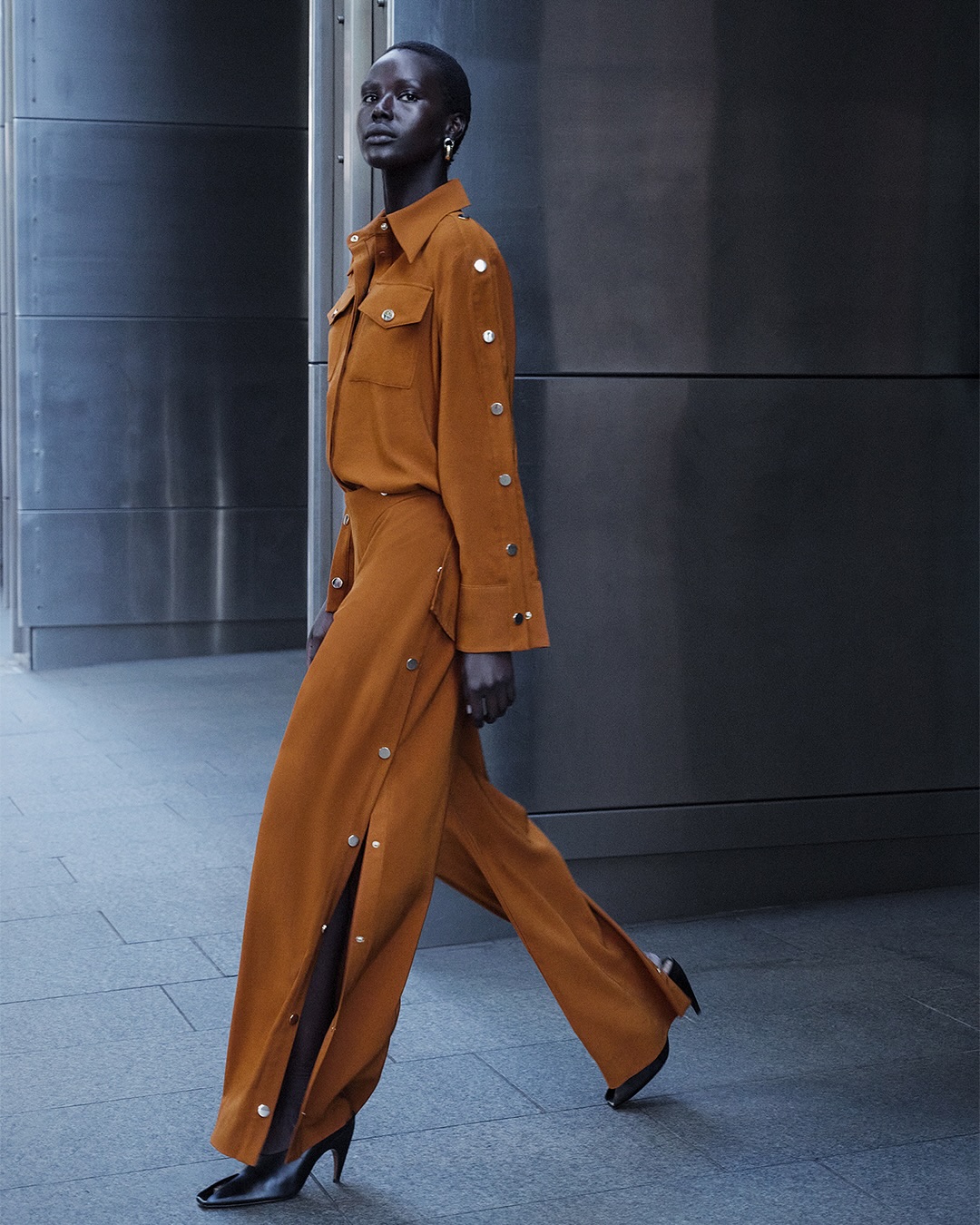 A dark-skinned model poses in a bronze-colored two-piece outfit, featuring a button-down shirt and wide-legged pants with side slits and heels.