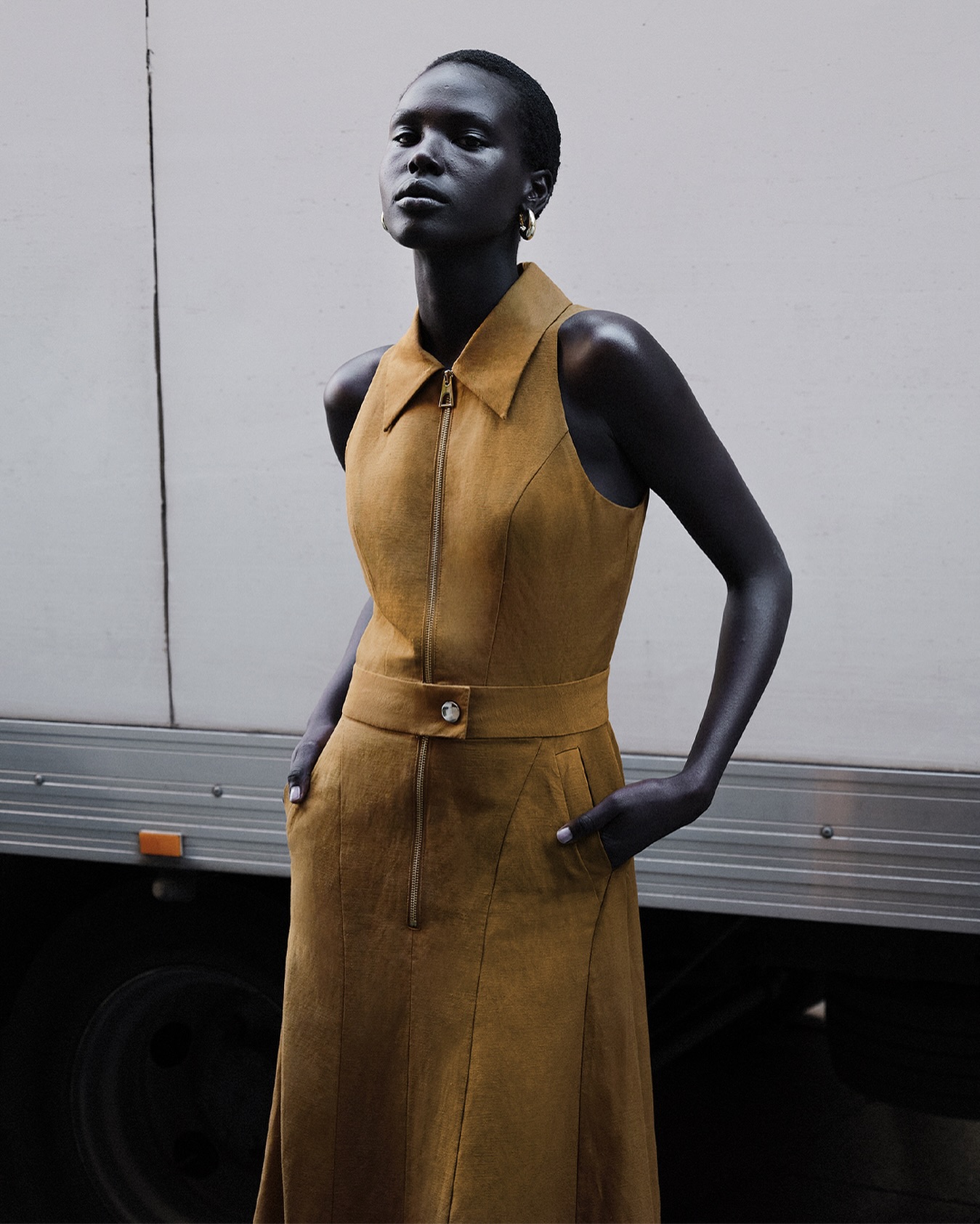 Woman in brown sleeveless dress with front zipper and belt posing near a trailer, showcasing fashion and editorial style with neutral tones.