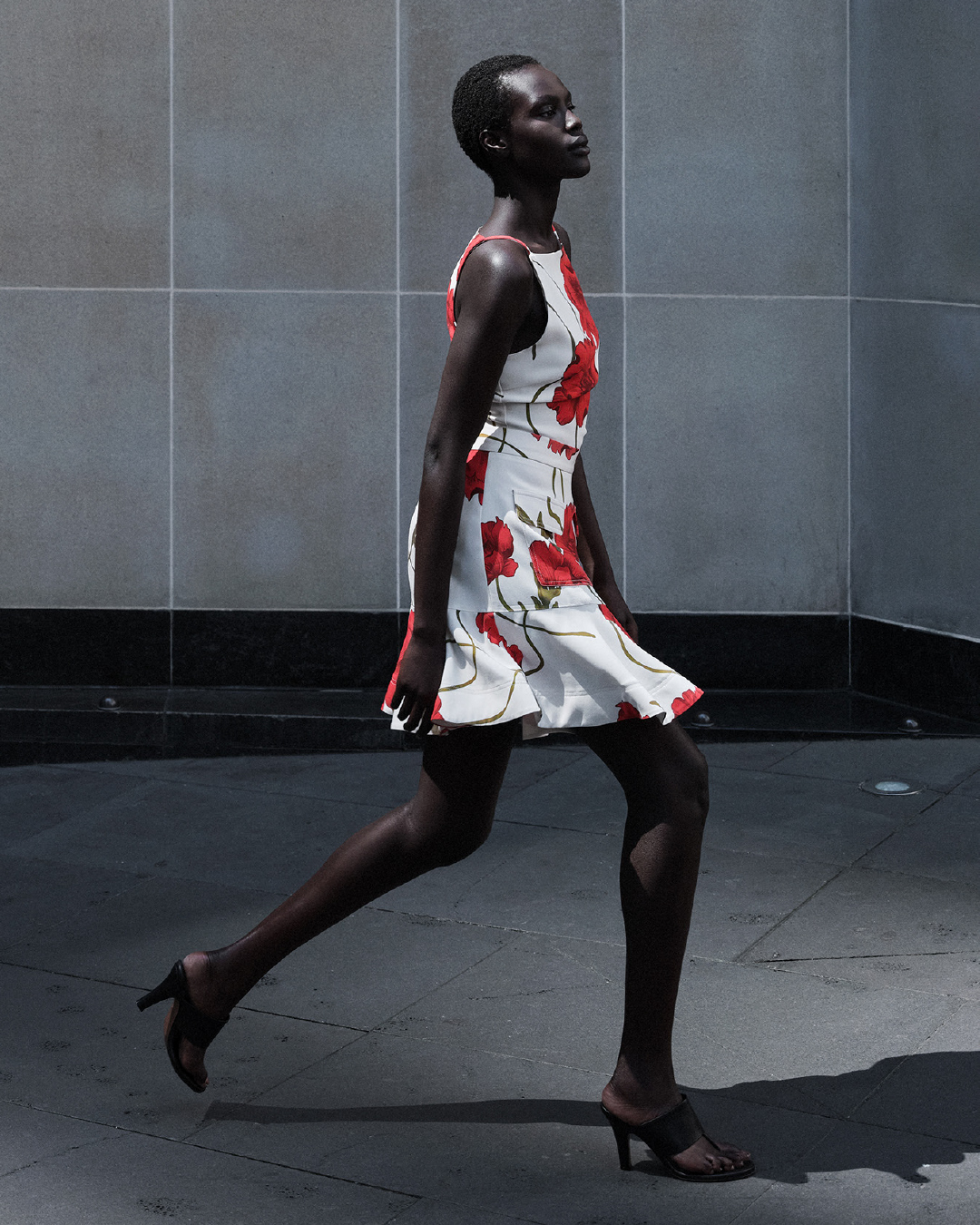 A dark-skinned adult female strides confidently, wearing a red and white floral print dress and heels, against a neutral urban backdrop.