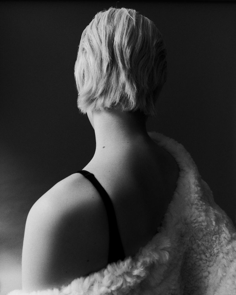Back view of a female figure with short blonde hair, wearing a black top and a white fur coat against a dark grey background.
