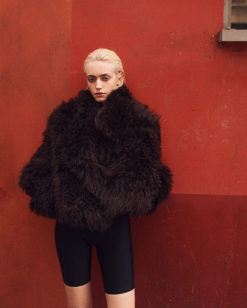 Woman in a fuzzy dark coat and black shorts poses against a red wall, fashion portrait, studio lighting.