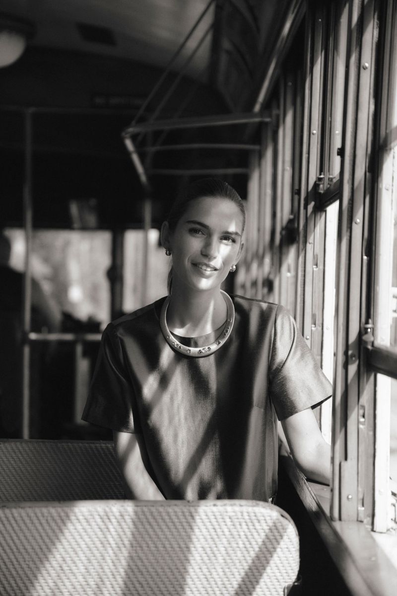 Monochrome portrait of a smiling woman with jewelry inside a vehicle with sunlight streaming through the windows and casting shadows.