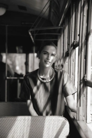 Monochrome portrait of a smiling woman with jewelry inside a vehicle with sunlight streaming through the windows and casting shadows.