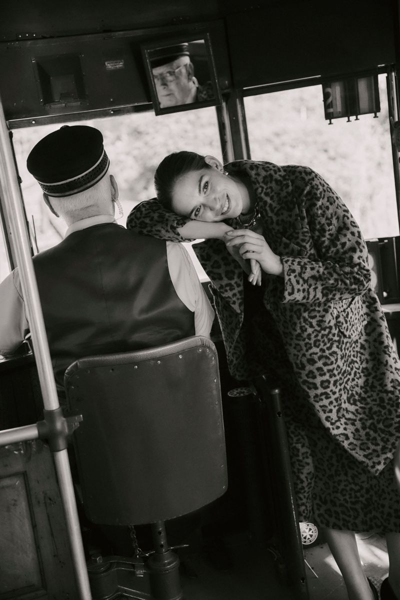 Black and white portrait of a smiling woman in a leopard print coat, leaning against the seat of a bus with a uniformed driver.
