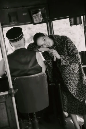 Black and white portrait of a smiling woman in a leopard print coat, leaning against the seat of a bus with a uniformed driver.
