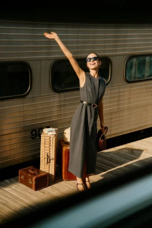 A fashionable woman waves goodbye at a train station in a sleeveless grey dress, sunglasses, and black accessories on a sunny day.