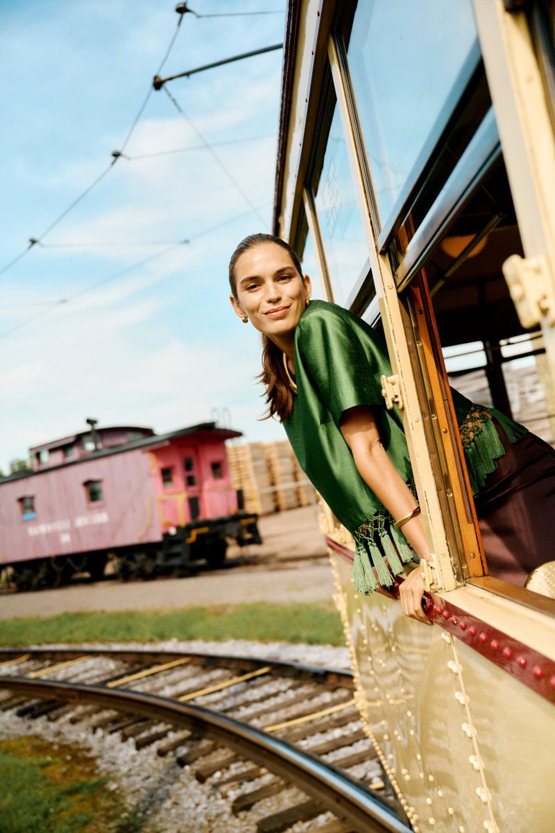 An adult woman smiles from the window of a vintage train car, wearing a stylish green top with tassels, railroad tracks visible.