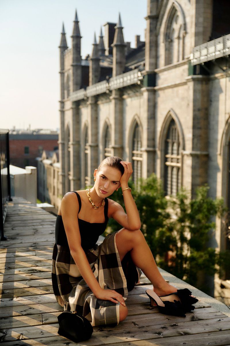 Elegant woman in a black tank top and plaid skirt sits on a wooden surface, posing near stone buildings with arched windows in daylight.