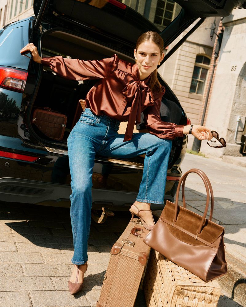 Woman in a satin blouse and jeans sits in the open trunk of a black car near a brown suitcase and leather handbag in an urban setting.