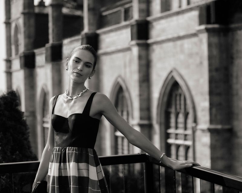 A woman poses in a black top and plaid skirt with a stone building backdrop, wearing jewelry and bracelet. The black and white image is stylish.