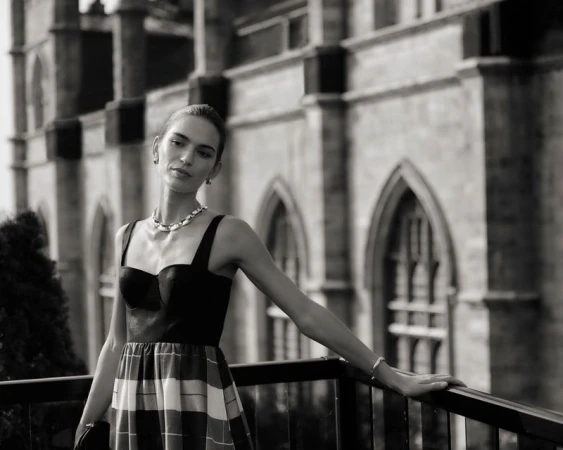 A woman poses in a black top and plaid skirt with a stone building backdrop, wearing jewelry and bracelet. The black and white image is stylish.