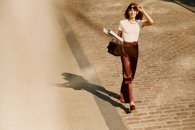 A smiling woman in stylish brown pants and a white top walks on a brick road, holding documents and a bag, accessorized with sunglasses.