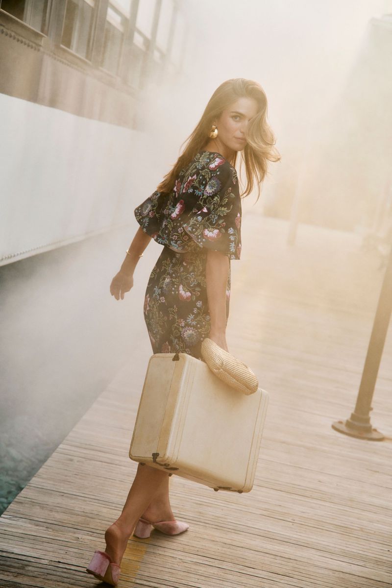 Woman in floral dress walks on a dock with a vintage suitcase in a hazy environment. Blonde hair, stylish earrings, and pink heels.