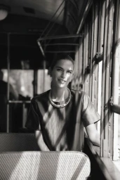 Model seated inside a vintage transit car wearing a dark top and statement necklace, lit by dramatic window light.
