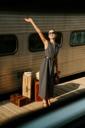 Smiling woman in a grey dress with vintage suitcases standing on a platform beside a train.