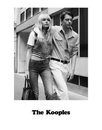 Black and white fashion shot of a couple in a city: a blonde woman in a varsity jacket and jeans with a bag, and a man in a shirt and white pants.