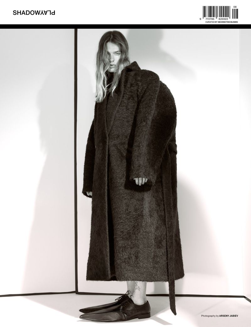 Fashion editorial shot of a woman with wavy blonde hair wearing an oversized, shaggy black coat and dress shoes in a bright white studio.