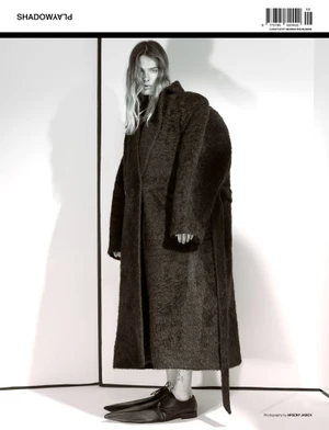 Model wearing a long dark fuzzy coat and oversized shoes standing against a plain white studio background