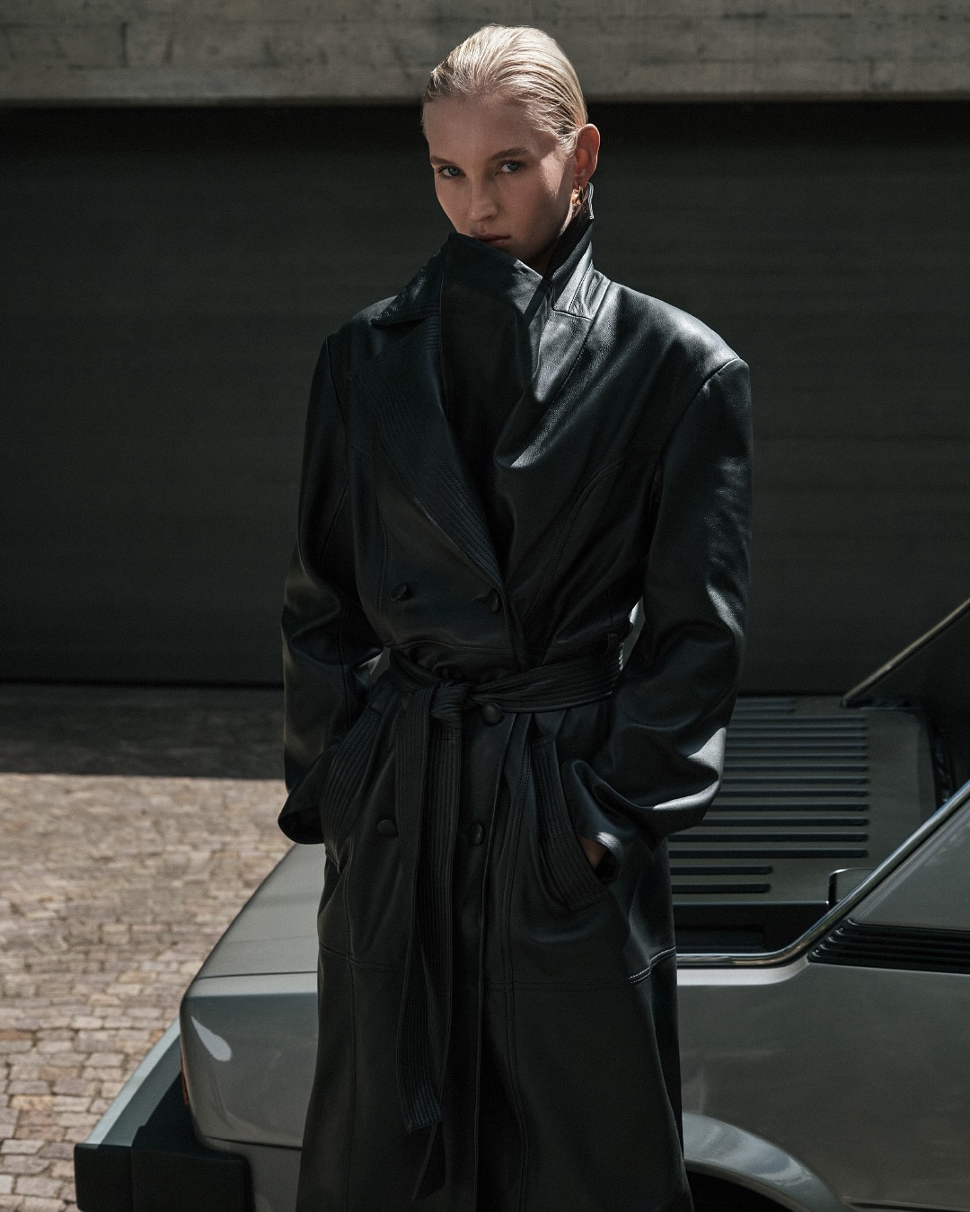 Adult female fashion model in black leather trench coat stands in front of a gray vehicle and closed gray metal door in outdoor shot.