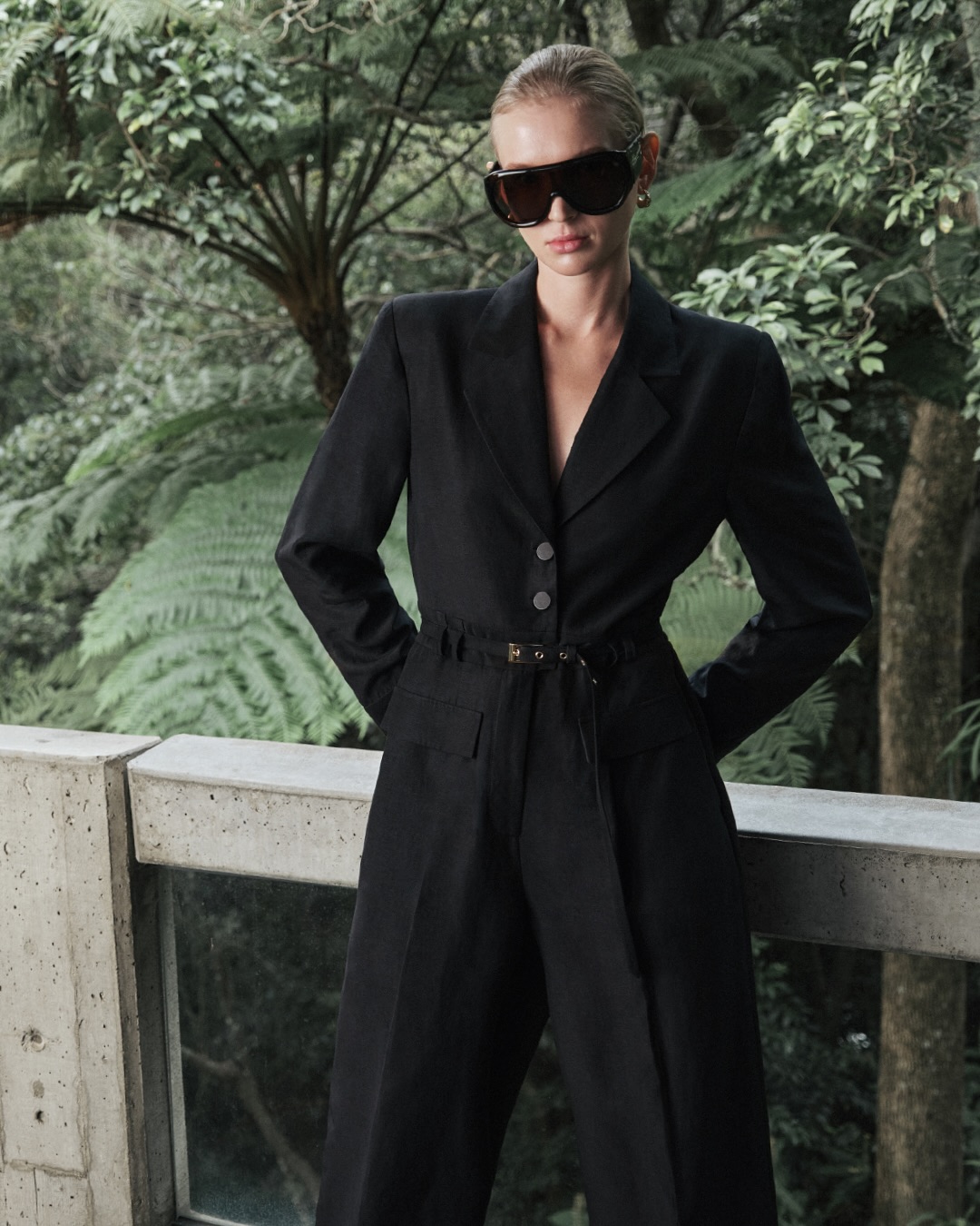 A blonde woman in a black suit and sunglasses strikes a pose outdoors against a backdrop of lush green ferns and foliage.