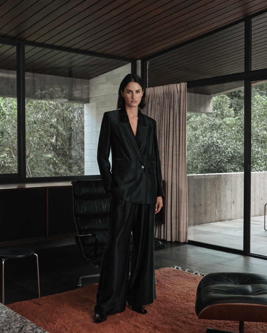 Indoor portrait of a woman wearing a black suit, standing in a modern architectural space with large windows, neutral tones, and natural light.