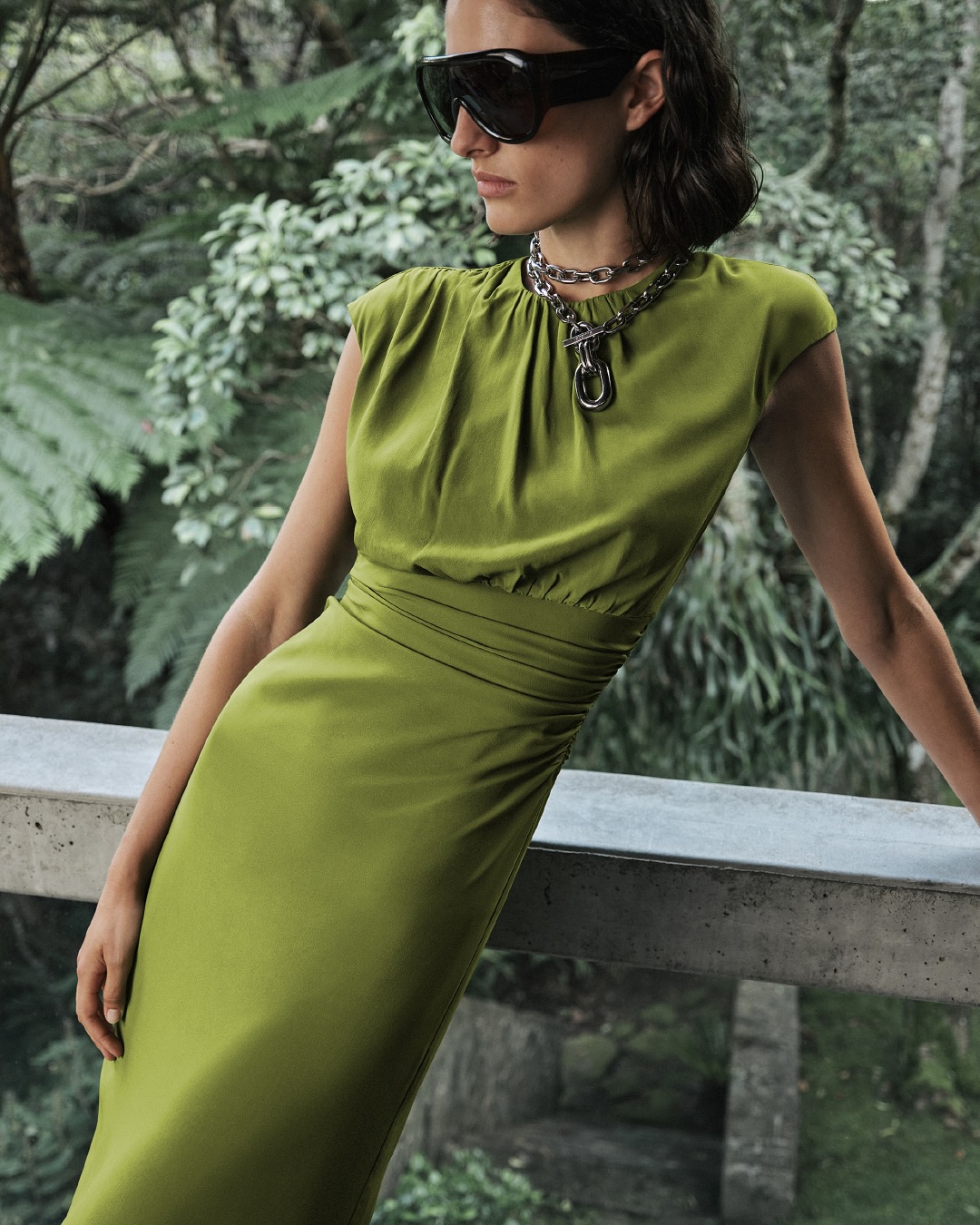 Woman in green dress and oversized sunglasses poses outdoors near lush foliage, showcasing fashion and elegant style.