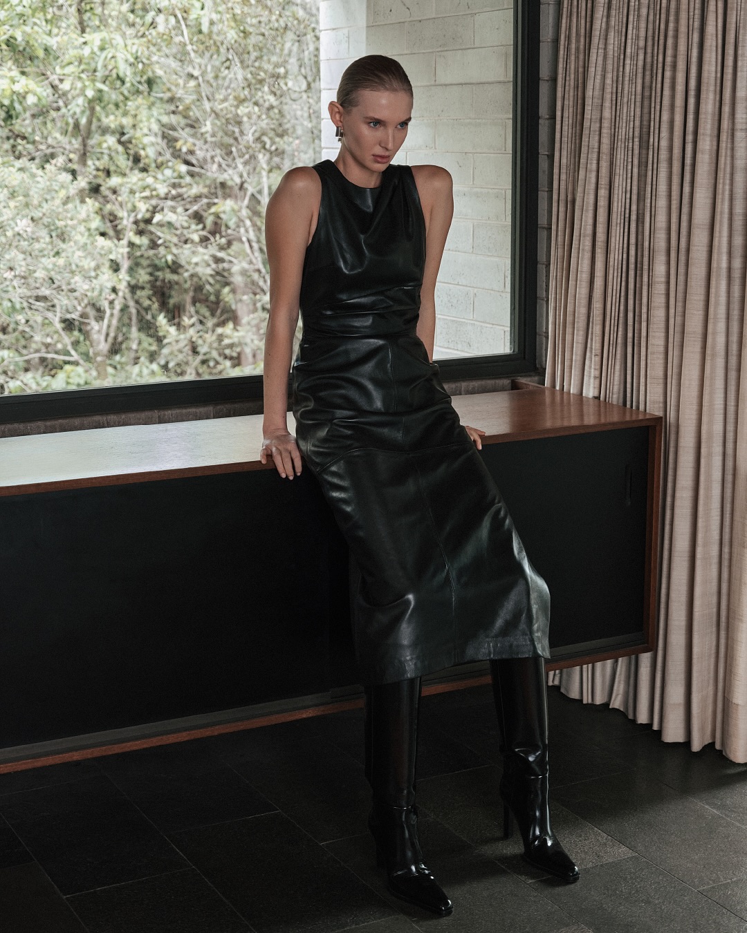 An adult woman wearing a black leather dress and black boots is sitting on a low console in a light-filled indoor environment.