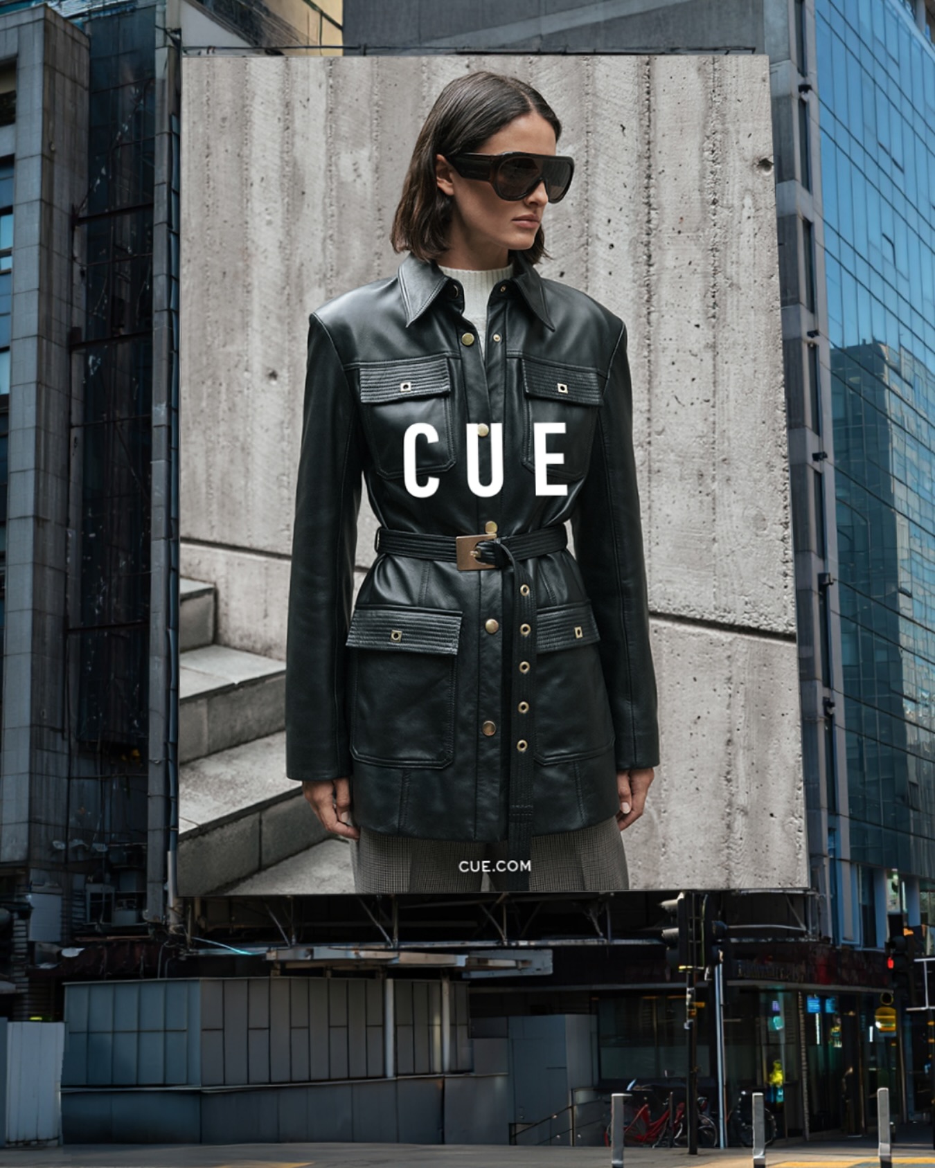 Fashion billboard featuring a woman wearing a leather jacket, sunglasses, and a neutral outfit, standing in front of a concrete wall.