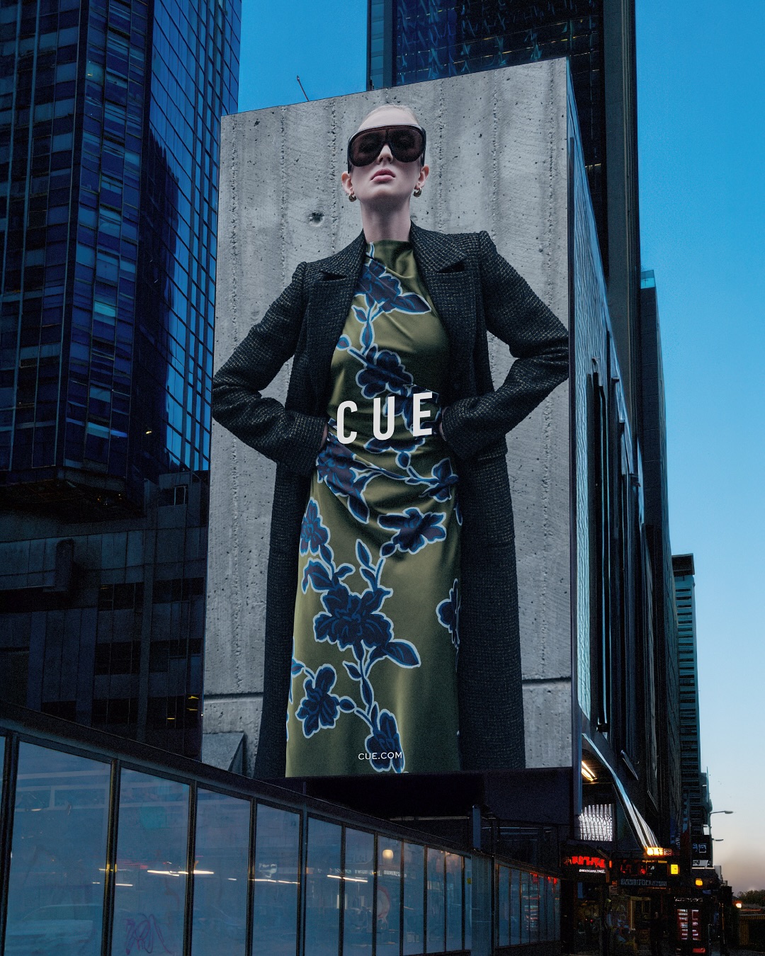 Outdoor fashion billboard featuring a stylish adult female model in a dress and coat with modern architecture and urban setting.