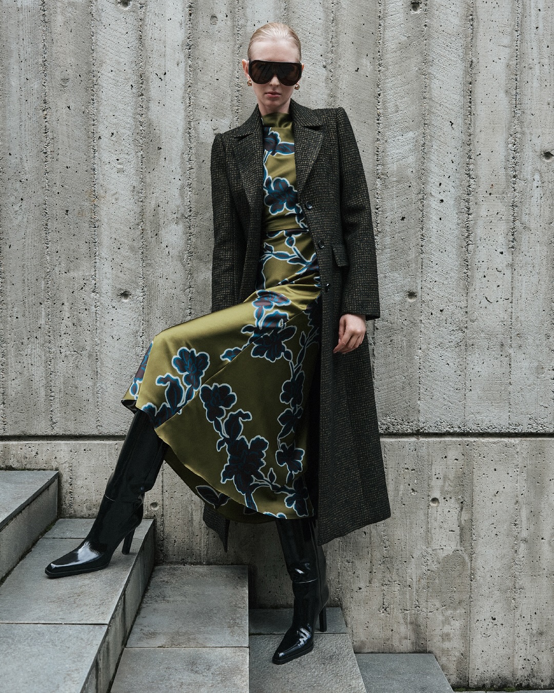 A woman poses on concrete stairs in a floral dress, dark sunglasses, coat, and high boots, against a concrete wall backdrop.