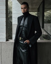 Model in a black leather vest, turtleneck, and long coat posing against a concrete architectural background.