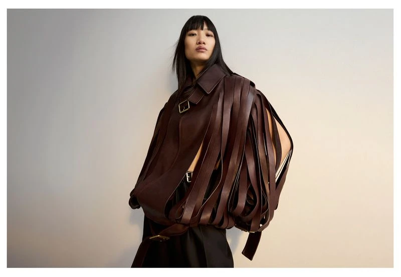 A fashion portrait features an Asian woman with dark hair in a brown leather jacket with fringe detail, against a simple neutral background.