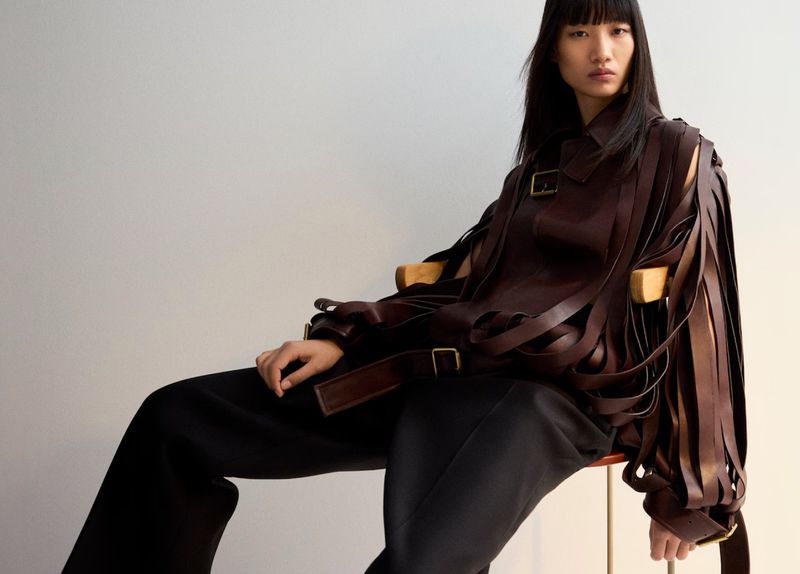 An Asian woman sits in a chair wearing a leather jacket with buckle straps, black pants, with a neutral studio backdrop.