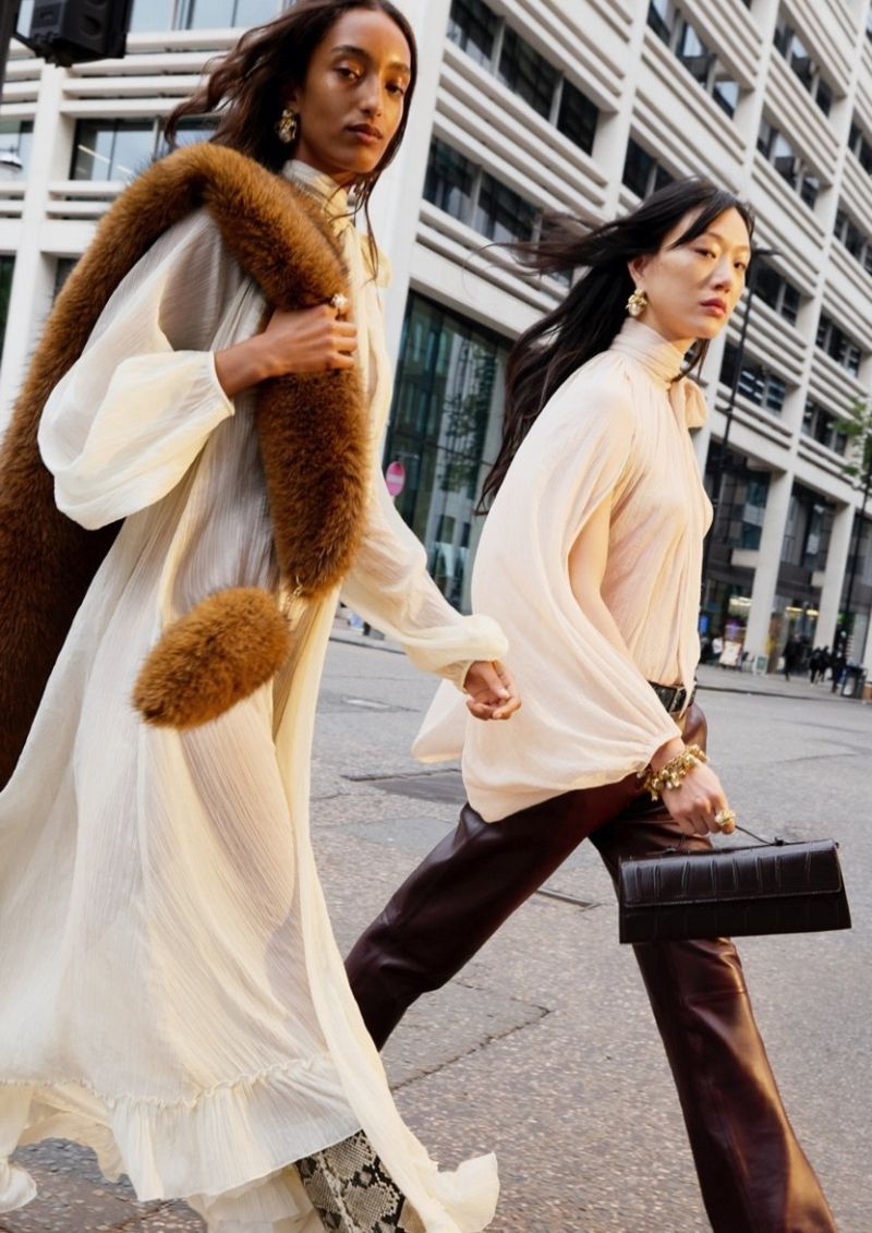 Two women in fashionable outfits stroll on a city street, one in a cream dress and the other in a beige top and brown pants.