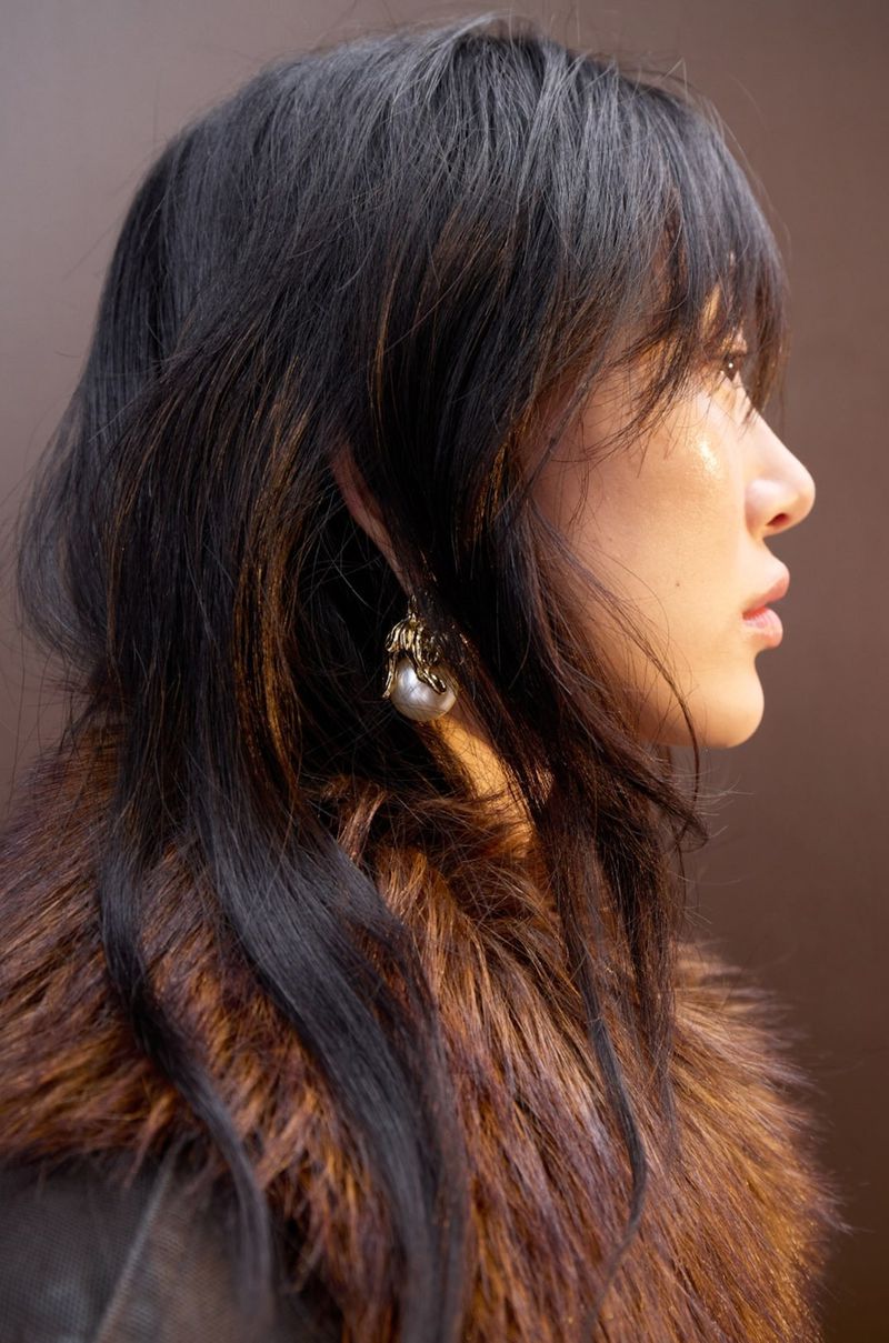 Profile close-up of model Sora Choi wearing a brown faux fur collar and a pearl drop earring against a warm brown backdrop.