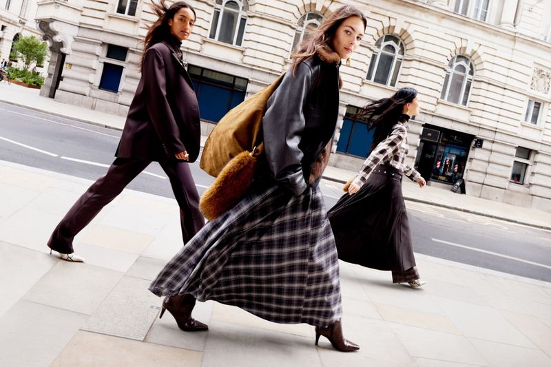 Three women in fashionable fall outfits walk on a city sidewalk. One woman carries a shoulder bag, all wear heeled boots.