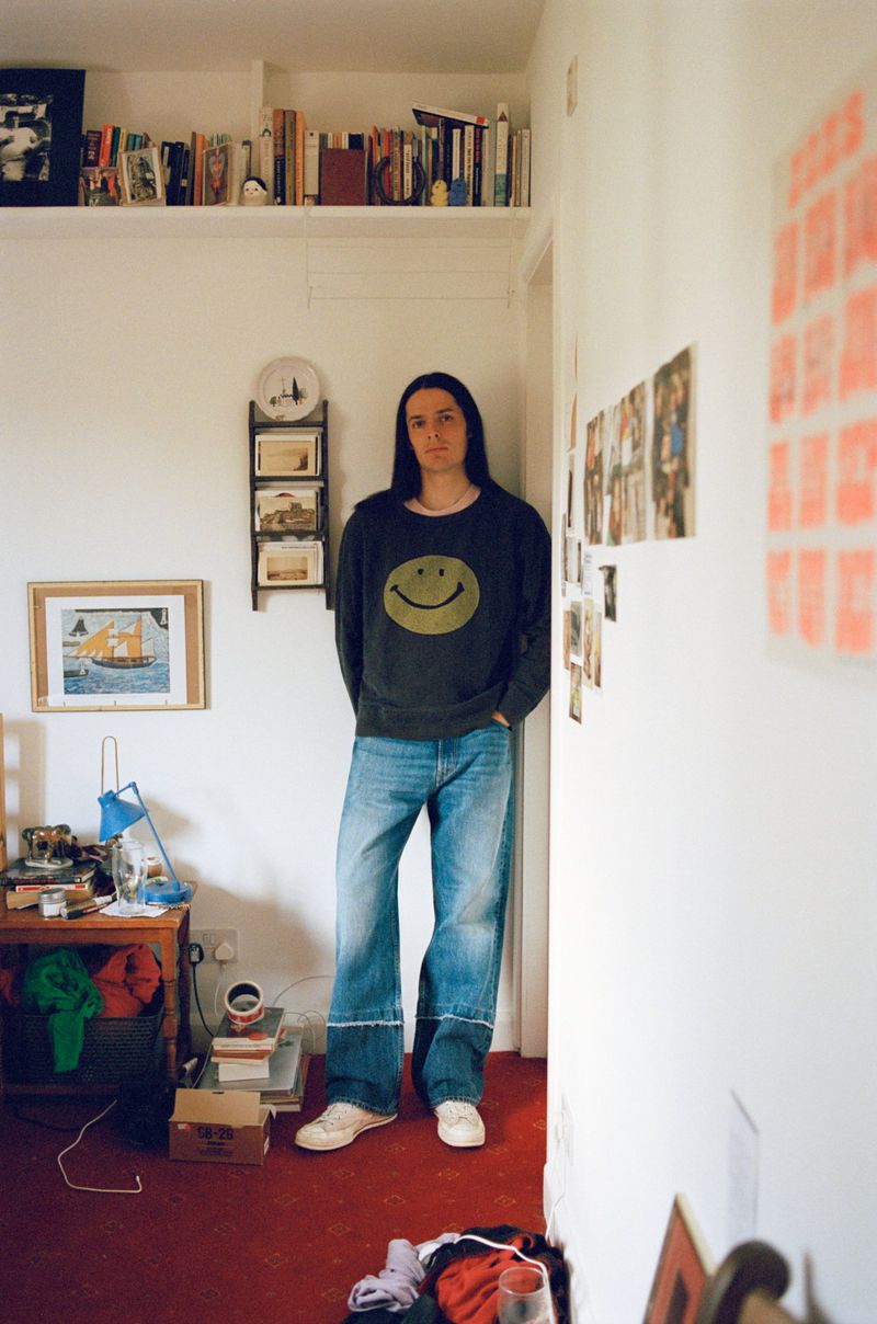 Indoor portrait of a young adult with long hair wearing a smiley-face sweatshirt and jeans, standing in a room with wall decorations and books.