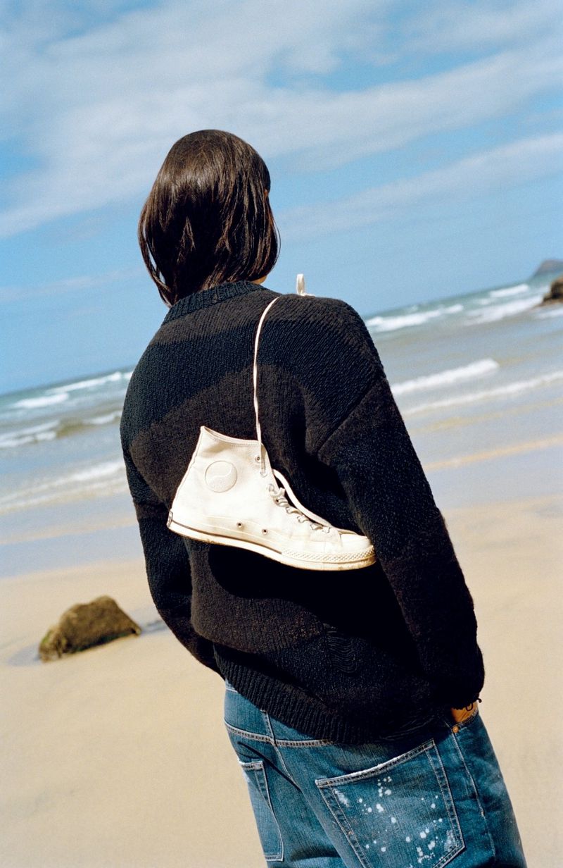Person from behind on beach with blue jeans, black sweater and a sneaker hanging from their shoulder with ocean in background.