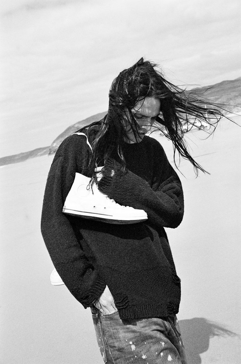 Monochrome image of a person with long dark hair wearing a sweater holding shoes on a beach with water and mountains in background.