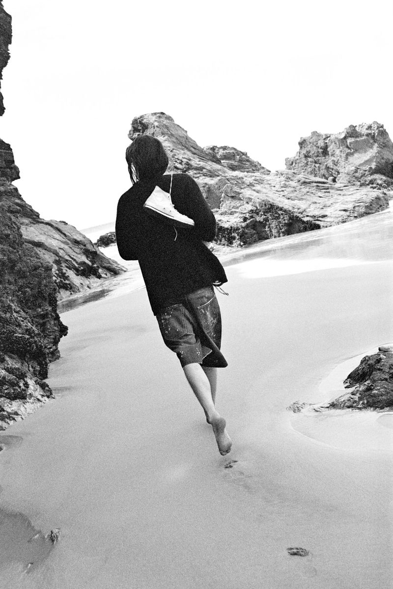 Back view of a barefoot adult running on a sandy beach, carrying a book; monochrome image featuring rocky cliffs and ocean.
