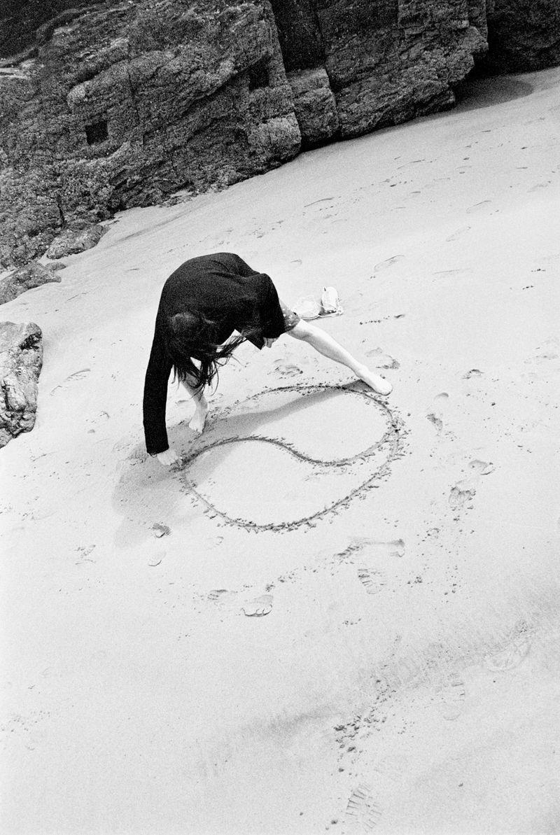 Woman in black dress drawing yin and yang symbol in the sand on a beach with rock formations in the background in black and white.