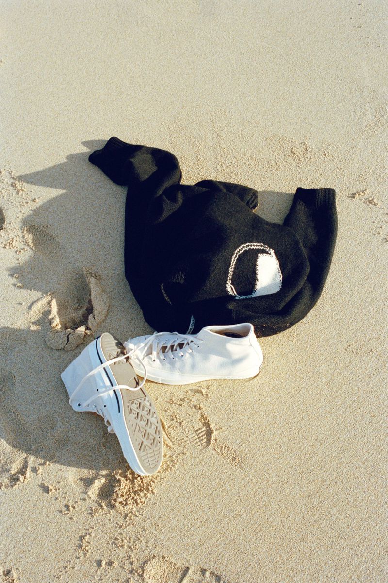 A black hooded sweatshirt and pair of white tennis shoes sit on light tan sand in bright sunlight, casting soft shadows near the beach.