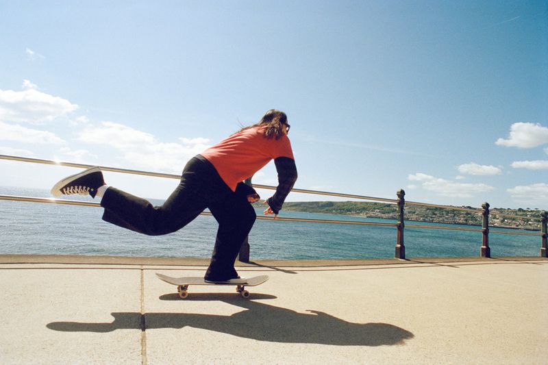 A skateboarder rides along a concrete path with a metal railing, ocean, and distant cityscape under a blue sky with scattered clouds.