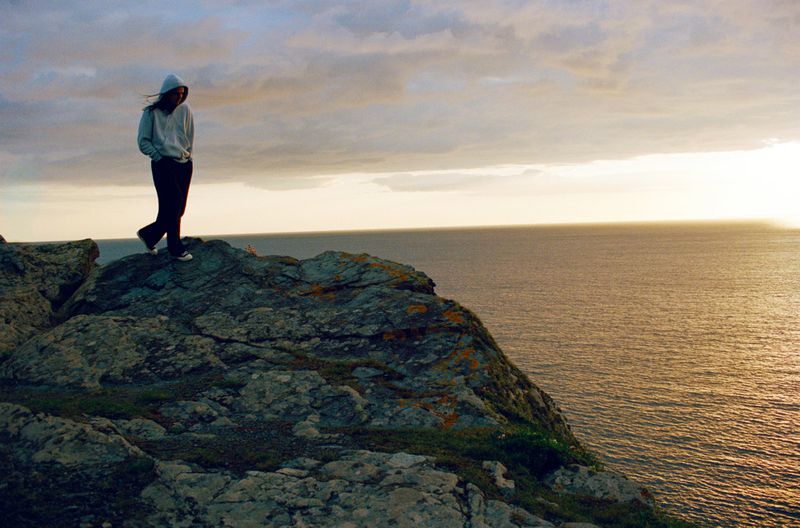 Woman in casual clothing walking on a rocky outcrop by the sea at sunset, with golden sunlight on the water and a cloudy sky.