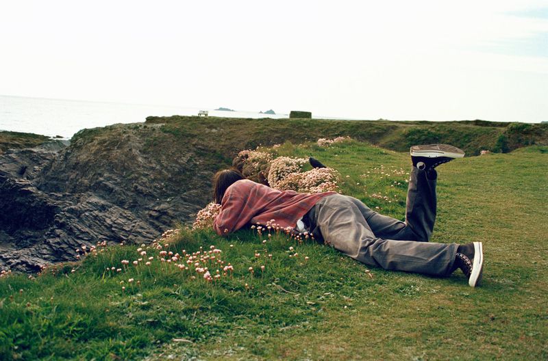 A person lies on a grassy hillside with wildflowers overlooking the ocean and rocky coastline under a clear sky in a scenic outdoor setting.