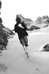 Model walking barefoot on a sandy beach carrying a white sneaker over their shoulder in a black and white fashion campaign image